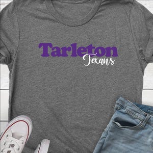 May include: A gray t-shirt with the words "Tarleton Texans" in purple and white. The shirt is paired with blue jeans and white sneakers.