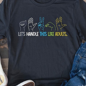 May include: A dark gray t-shirt with a graphic of five hands in the American Sign Language alphabet spelling the word "adult". The text "Let's handle this like adults." is printed below the graphic.