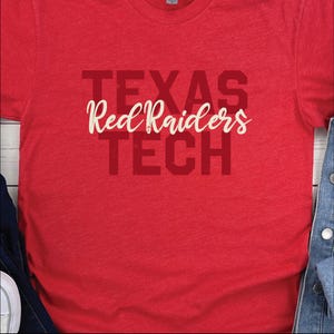 May include: A heather red short-sleeved t-shirt featuring the text TEXAS TECH in a bold, dark red block font. Overlaid in the center is the phrase Red Raiders written in a white, cursive script font, creating a layered graphic design.