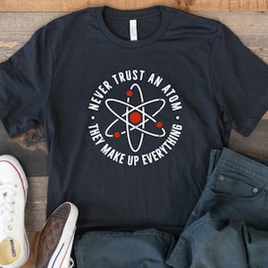 May include: A black t-shirt with a white circle graphic featuring an atom and the text "Never trust an atom. They make up everything."