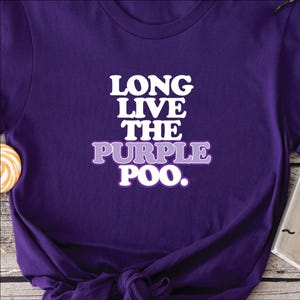 May include: A purple t-shirt with the text "LONG LIVE THE PURPLE POO" in white and purple lettering. The shirt is knotted at the bottom. A lollipop and a small black object are visible in the background.