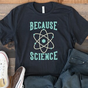 May include: A black t-shirt with the text "Because Science" in teal blue lettering. A stylized teal blue atom graphic is centered below the text.