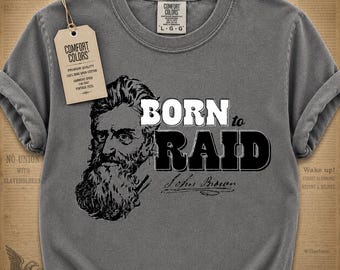 Pode incluir: Camiseta cinza Comfort Colors com a frase "BORN to RAID" e um retrato de John Brown. A camisa tem um toque vintage e uma etiqueta que diz "Comfort Colors".