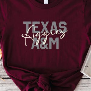 May include: Maroon t-shirt with the text "TEXAS Aggies A&M" in gray and white. The shirt is knotted at the bottom. The shirt is made of a soft material.
