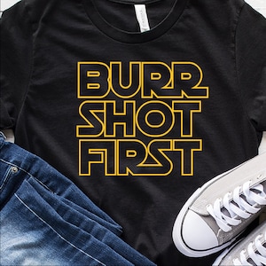 Pode incluir: Camiseta preta com o texto "Burr Shot First" em letras amarelas com contorno.