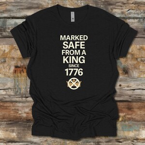 May include: Black t-shirt with the text "MARKED SAFE FROM A KING SINCE 1776" in a cream-colored font. The design includes a crown and emblem. The shirt is displayed on a wooden surface.