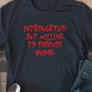 May include: Dark navy blue t-shirt with the red text "INTROVERTED BUT WILLING TO DISCUSS ANIME". The shirt is folded, with a pair of blue jeans and a white shoe visible in the image. The text is in a bold, stylized font.