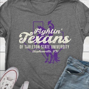 May include: A heather gray t-shirt featuring the text "Fightin' Texans" in a stylized font, with a purple outline of the state of Texas. Below the text is "OF TARLETON STATE UNIVERSITY Stephenville, TX". A pair of white sneakers and blue jeans are visible.