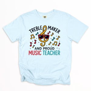 May include: Light blue t-shirt with the text "TREBLE MAKER AND PROUD MUSIC TEACHER". The design features a yellow treble clef wearing heart-shaped sunglasses, surrounded by colorful musical notes.