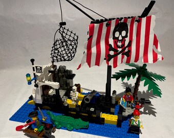 Könnte beinhalten: Ein Lego Piratenschiff-Set mit einer rot-weiß gestreiften Piratenflagge, einem schwarzen Totenkopf mit Knochen, einem Netz, einer Palme und mehreren Lego-Minifiguren.