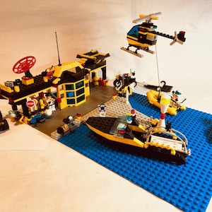 Puede incluir: Un set de Lego City que representa una escena portuaria con una estación, un barco, un helicóptero y otros vehículos. Los edificios son negros y amarillos, con un radar rojo. El barco y el helicóptero son amarillos y negros. La escena está sobre una placa base azul.