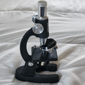 Puede incluir: Un microscopio negro vintage con perillas de ajuste plateadas y un ocular cromado. El microscopio tiene una base robusta y un brazo curvo, típico de los instrumentos científicos antiguos. Está diseñado para uso educativo o aficionado.