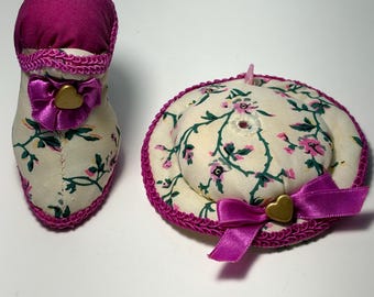 May include: A fabric shoe and hat set. The shoe is cream-colored with a floral pattern, a fuchsia top, and a fuchsia trim. The hat is cream-colored with a floral pattern, a fuchsia trim, and a fuchsia bow with a gold heart.