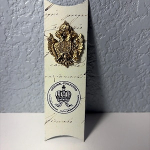 May include: A gold-colored metal pin in the shape of a double-headed eagle, mounted on a cream-colored card with script and a circular logo that reads "ORIGINAL COLLECTION". The pin is highly detailed and ornate.