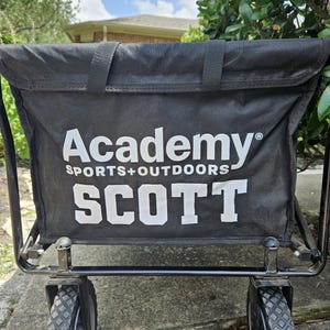 May include: Black utility cart with the text "Academy SPORTS+OUTDOORS SCOTT" in white. The cart has a black fabric bag, a black metal frame, and two black and gray wheels. The cart is designed for outdoor use.