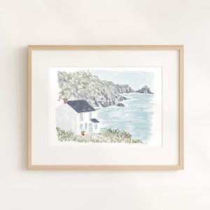 May include: Framed watercolor print of a white house with a dark blue roof, situated on a cliff overlooking the ocean. The artwork features a light blue sky, rocky cliffs, and a serene coastal landscape. The frame is a light wood color.