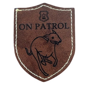 Puede incluir: Un parche de cuero marrón en forma de escudo con costuras blancas. El parche presenta el texto "ON PATROL" sobre una ilustración de un perro corriendo. Una pequeña insignia está en la parte superior.