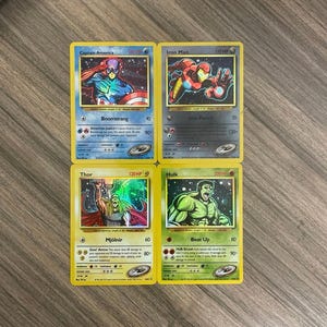 May include: Four collectible trading cards featuring Marvel superheroes: Captain America, Iron Man, Thor, and Hulk. Each card has a unique design with character illustrations, names, and special abilities. The cards are on a wooden surface.