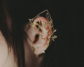 May include: A gold ear cuff with a delicate branch design. The cuff is worn on the upper part of the ear and features small leaves.