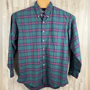 Puede incluir: Camisa a cuadros verdes, rojos y azules. La camisa de manga larga tiene cuello abotonado, bolsillo en el pecho y botones blancos. La camisa está colgada de una percha de madera sobre un fondo de madera clara.