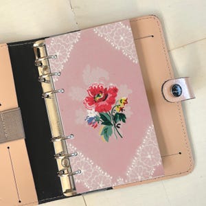 Op de afbeelding: Een roze notitieboekinlegvel met een bloemenpatroon en een witte kantzoom. De inlegvel is ontworpen om te passen in een ringband van standaardformaat.