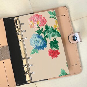 Op de afbeelding: Een crèmekleurige planner met een bloemenontwerp met roze, blauwe en rode bloemen. De planner is open en heeft een zilveren ringband.
