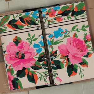 Op de afbeelding: Een witte planner met een bloemenontwerp met roze, blauwe, groene en oranje bloemen. De planner heeft een zilveren binder met zes ringen.