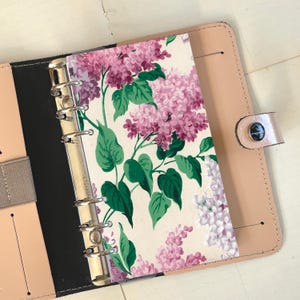 Op de afbeelding: Een zwarte en beige leren map met een bloemenpatroon insert. De insert heeft een witte achtergrond met roze en groene lila bloemen.