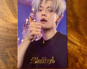 May include: A rectangular photo card featuring a person with light-coloured hair giving a thumbs up. The person wears a black shirt against a purple background with a blurred chandelier. The gold text "Stuckissed" is printed at the bottom centre.