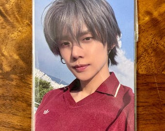 May include: A rectangular collectible photo card featuring a person with short, layered, ash-brown hair. The person wears a dark red V-neck shirt with a small white logo on the chest and a white trim on the collar. The background shows a blue sky.