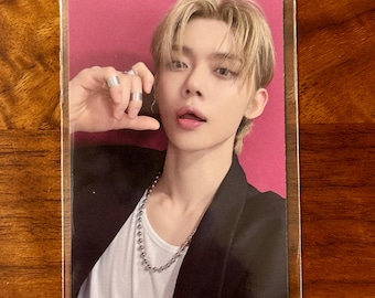 May include: A rectangular trading card featuring a portrait of a person with blonde hair, wearing a black blazer over a white shirt and a silver ball chain necklace. The person has silver rings on their fingers and is set against a solid pink background.