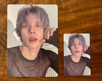 May include: Two rectangular photo cards featuring a portrait of a person with short, light purple hair. The person is wearing a black long-sleeved shirt with a sheer texture and has one hand raised near their face with their tongue slightly visible.