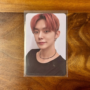 May include: A rectangular trading card featuring a portrait of a person with reddish-brown hair, wearing a black crew-neck shirt and a thin black beaded necklace. The person has small hoop earrings and is positioned against a plain white background.