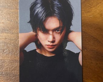 May include: A rectangular collectible photo card featuring a portrait of a person with dark, layered hair and a black t-shirt. The individual has their hands positioned behind their head, looking directly forward against a solid grey background.