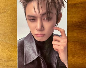 May include: A rectangular collectible photo card featuring a portrait of a person with light brown hair, wearing a dark brown leather jacket over a black turtleneck. The person has one finger touching their cheek and is looking directly forward.