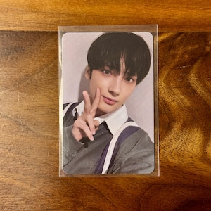 May include: A rectangular collectible photo card encased in a clear plastic sleeve featuring a portrait of a young person with dark hair, wearing a dark gray sweater over a white collared shirt, making a peace sign gesture with their right hand.