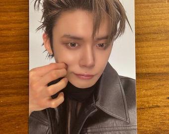 May include: A rectangular collectible photo card featuring a portrait of a person with brown hair styled in a messy, layered cut. The individual wears a black leather jacket with a visible collar and a black shirt underneath, while touching their cheek.