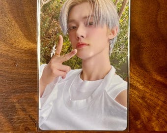 May include: A rectangular trading card featuring a portrait of a person with light-coloured hair, wearing a white sleeveless top with a layered neckline. The person is making a peace sign with one hand, which is adorned with a small white ring.