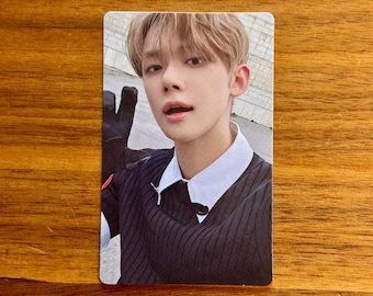 May include: A rectangular trading card featuring a portrait of a young person with light brown hair. The individual wears a black ribbed sweater over a crisp white collared shirt. One hand, covered by a black glove, is raised near the face.