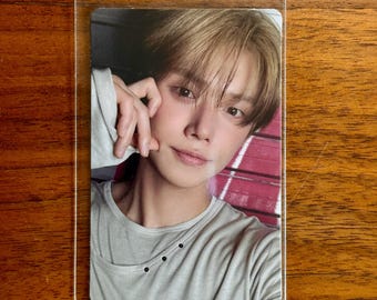 May include: A rectangular collectible photo card featuring a portrait of a person with light brown hair, wearing a light grey crew-neck shirt with three small black buttons near the neckline, enclosed in a clear protective plastic sleeve.