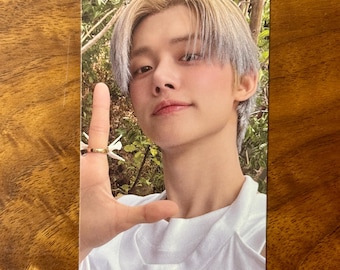 May include: A rectangular photo card featuring a person with light-colored hair, wearing a white shirt, and holding up one hand with a gold ring on the finger. The background shows green foliage and trees in a natural outdoor setting.