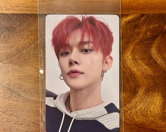 May include: A rectangular collectible photo card encased in a clear plastic sleeve featuring a portrait of a person with vibrant red hair, wearing a dark navy and white striped knit hooded sweater and small silver hoop earrings on both ears.