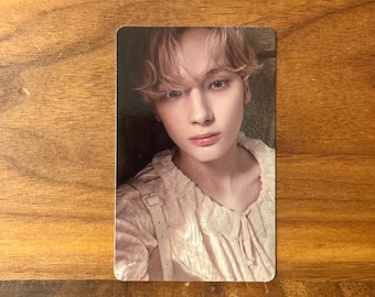 May include: A rectangular collectible photo card featuring a portrait of a person with light brown hair, wearing a pale pink long-sleeved shirt with a lace-trimmed collar and a vertical strap detail across the chest, set against a neutral background.