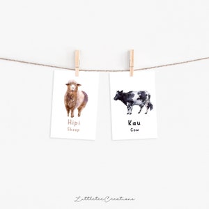 K&ouml;nnte beinhalten: Zwei Aquarell-Illustrationen von Nutztieren auf wei&szlig;en Karten, die an einer Schnur mit W&auml;scheklammern h&auml;ngen. Die Karte links zeigt ein braunes Schaf mit dem Text "Hipi Sheep". Die Karte rechts zeigt eine schwarz-wei&szlig;e Kuh mit dem Text "Kau Cow".