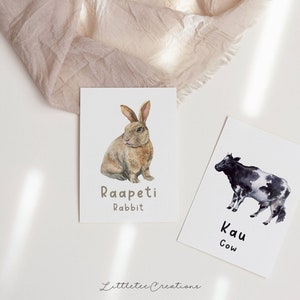 K&ouml;nnte beinhalten: Zwei Aquarell-Illustrationen von Nutztieren auf wei&szlig;en Karten. Die erste Karte zeigt ein braunes Kaninchen mit dem Text "Raapeti Rabbit". Die zweite Karte zeigt eine schwarz-wei&szlig;e Kuh mit dem Text "Kau Cow".