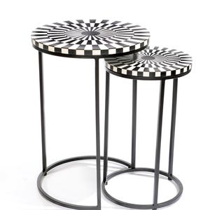 May include: Two nesting side tables with black metal frames. The round tabletops feature a black and white checkered pattern radiating from the centre. The tables have a modern design and are suitable for living rooms or bedrooms.