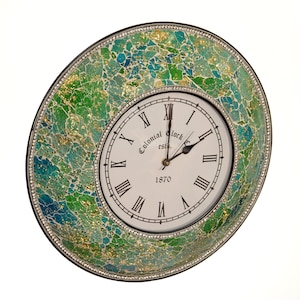 May include: A decorative wall clock with a mosaic-style surround in green, blue, and gold. The clock face is white with black Roman numerals and the text "Colonial Clock est. 1870". The bezel is trimmed with small crystals.
