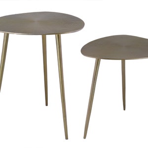 May include: A set of two gold metal side tables with a unique leaf-shaped top. The tables have a textured surface and thin, tapered legs.