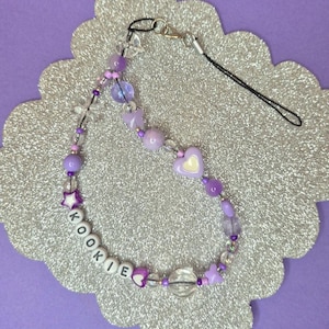 以下が含まれることがあります： 紫色のビーズの携帯電話チャーム。透明と紫色のビーズ、ハートと星の形をしたアクセント、白い文字ビーズで綴られた「KOOKIE」という名前が特徴です。黒いストラップとシルバーカラーの留め金が付いています。