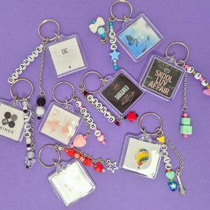 May include: A collection of clear square keychains with various designs and text, including "WINGS", "BE", and "SKOOL LUV AFFAIR". Each keychain has a silver ring and decorative beads. The background is a solid purple.